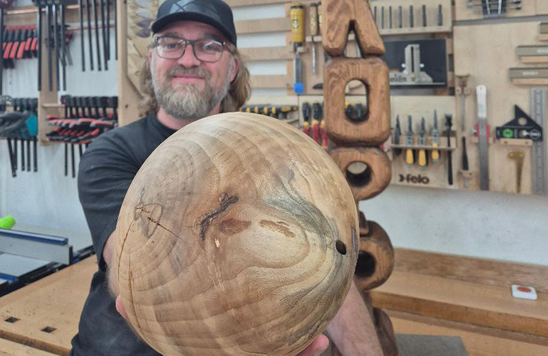 Winfried Engber holding out a large carved wooden sphere, its growth rings and bark inclusions visible