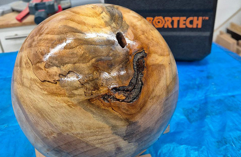 A polished wooden sphere with dark bark inclusions, resting on a bench beside an Arbortech case